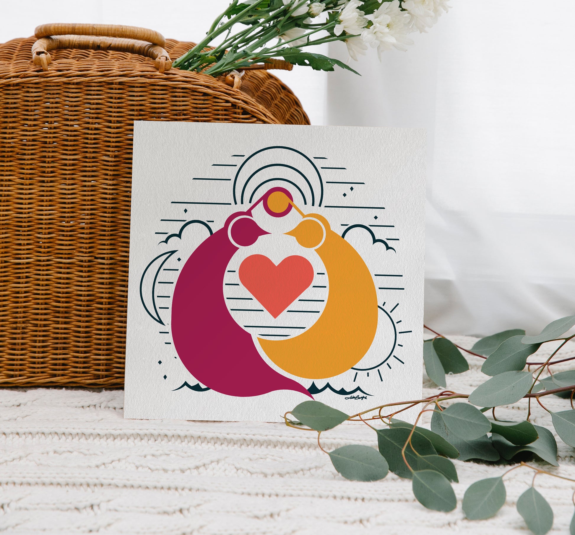 A 12"x12" print leaning against a wicker bag. Eucalyptus leaves are in the foreground. The illustrated print depicts two quails facing each other with their top knots overlapping. A heart fits in between them. There is a crescent moon, a sun, clouds, stars, and mountains in the background of the illustration.