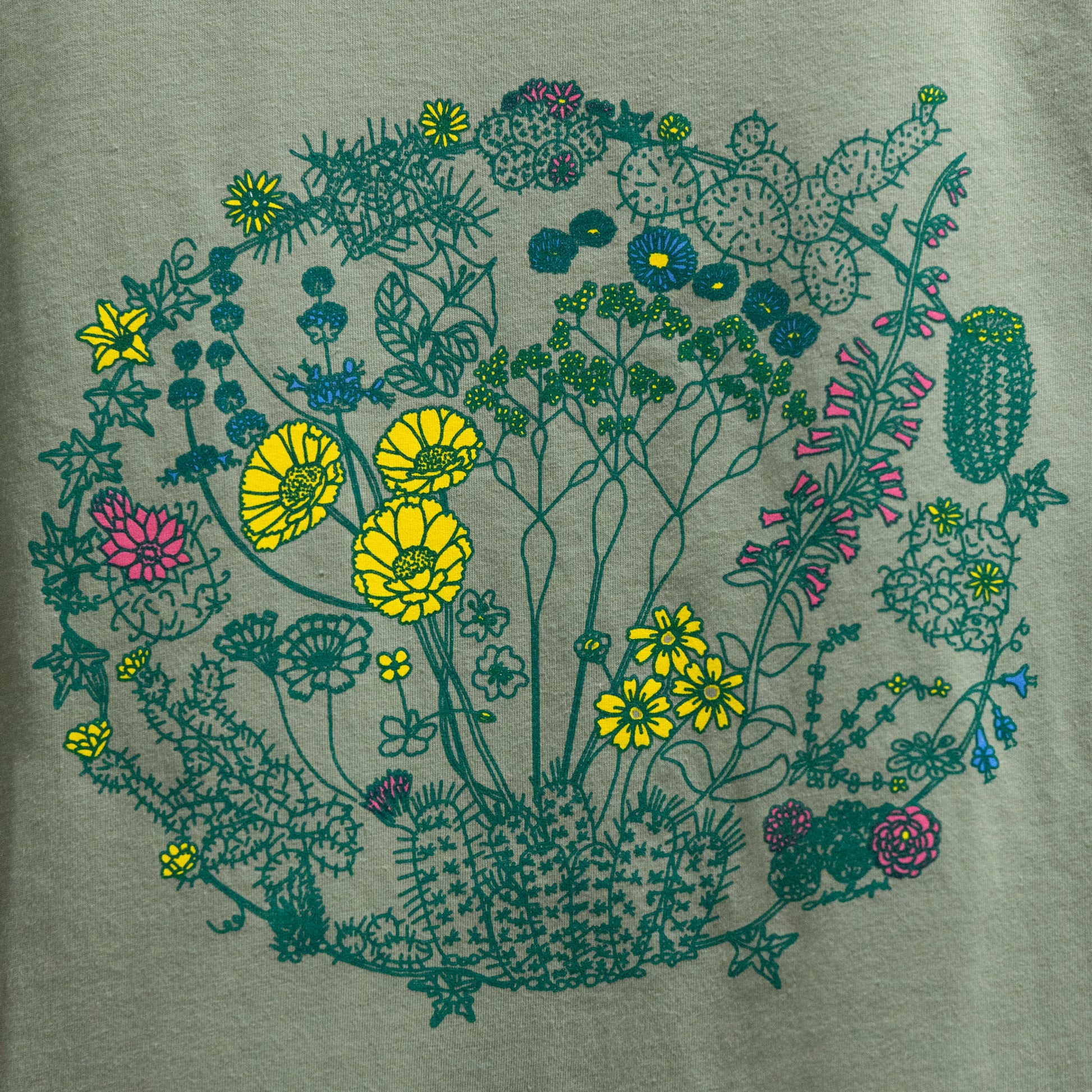 A close-up of a sage color t-shirt with an illustration of flowers on plants that can be found in the Mojave Desert and/or Joshua Tree National Park. The outlines of the plants are a dark green color. Some of the flowers are colored a bright yellow, some a bright pink, and some a bright blue. The composition is in a circular shape.