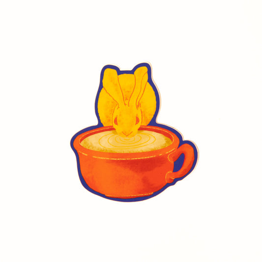 An illustrated drawing called Fika Bunny as a sticker. The illustration shows a yellow furred Desert Cottontail Rabbit taking a sip out of a large mug of coffee. The coffee mug is red in color and has a handle on the right side. The border of the sticker is a bright blue.