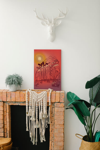 An illustrated print of Joshua Tree rocks and a few desert critters and plants. The rocks each have their own personality. Colors throughout resemble the beautiful desert sunsets. The print is resting against a white wall on top of a brick fireplace mantel. There is a macrame wall hanging from the top of the fireplace, two plants to the sides of the print, and a fake white deer head hangs above the print.