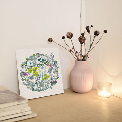 A 12x12 print rests against a white wall on top of a wood tabletop. There is a vace of dried flowers and a burning candle to the right of it and a stack of magazines to the left of it. The illustration is of flowers on plants that can be found in the Mojave Desert and/or Joshua Tree National Park. The outlines of the plants are a dark green color. Some of the flowers are colored a bright yellow, some a bright pink, and some a bright blue. The composition is in a circular shape.
