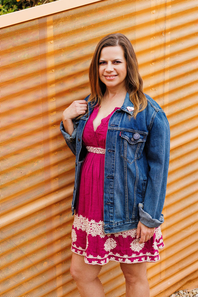 Lisa Burford wearing a denim jacket over a magenta dress with embroidered details. She is standing in front of a yellow mesh wall and has her right hand touching her shoulder.