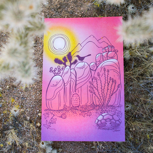 An illustrated print of Joshua Tree rocks and a few desert critters and plants. The rocks each have their own personality. Colors throughout resemble the beautiful desert sunsets. The print lays on the desert floor beneath a cholla cactus.