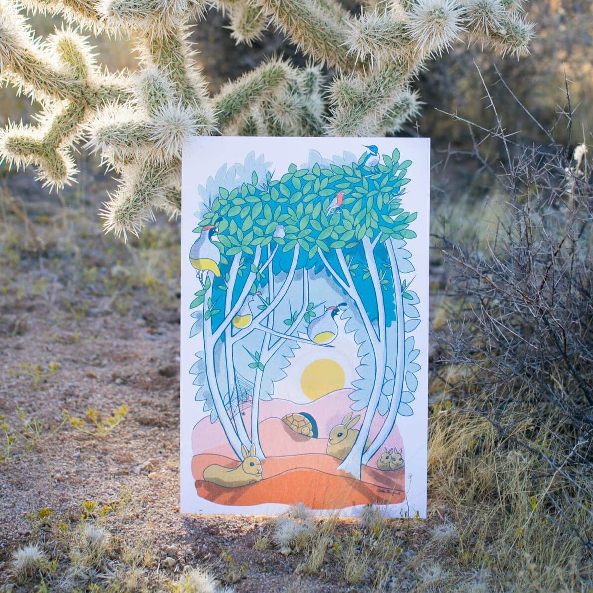 This 11"x17" print of animal characters found in a graythorn bush. Quail, cottontail bunnies, Western scrub jay, house finches, and a desert tortoise all hang out inside and underneath the branches of this bush. A sun can be seen setting in the background through an opening in the branches. The print rests against a cholla cactus.