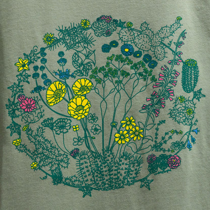 A close-up of a sage color t-shirt with an illustration of flowers on plants that can be found in the Mojave Desert and/or Joshua Tree National Park. The outlines of the plants are a dark green color. Some of the flowers are colored a bright yellow, some a bright pink, and some a bright blue. The composition is in a circular shape.