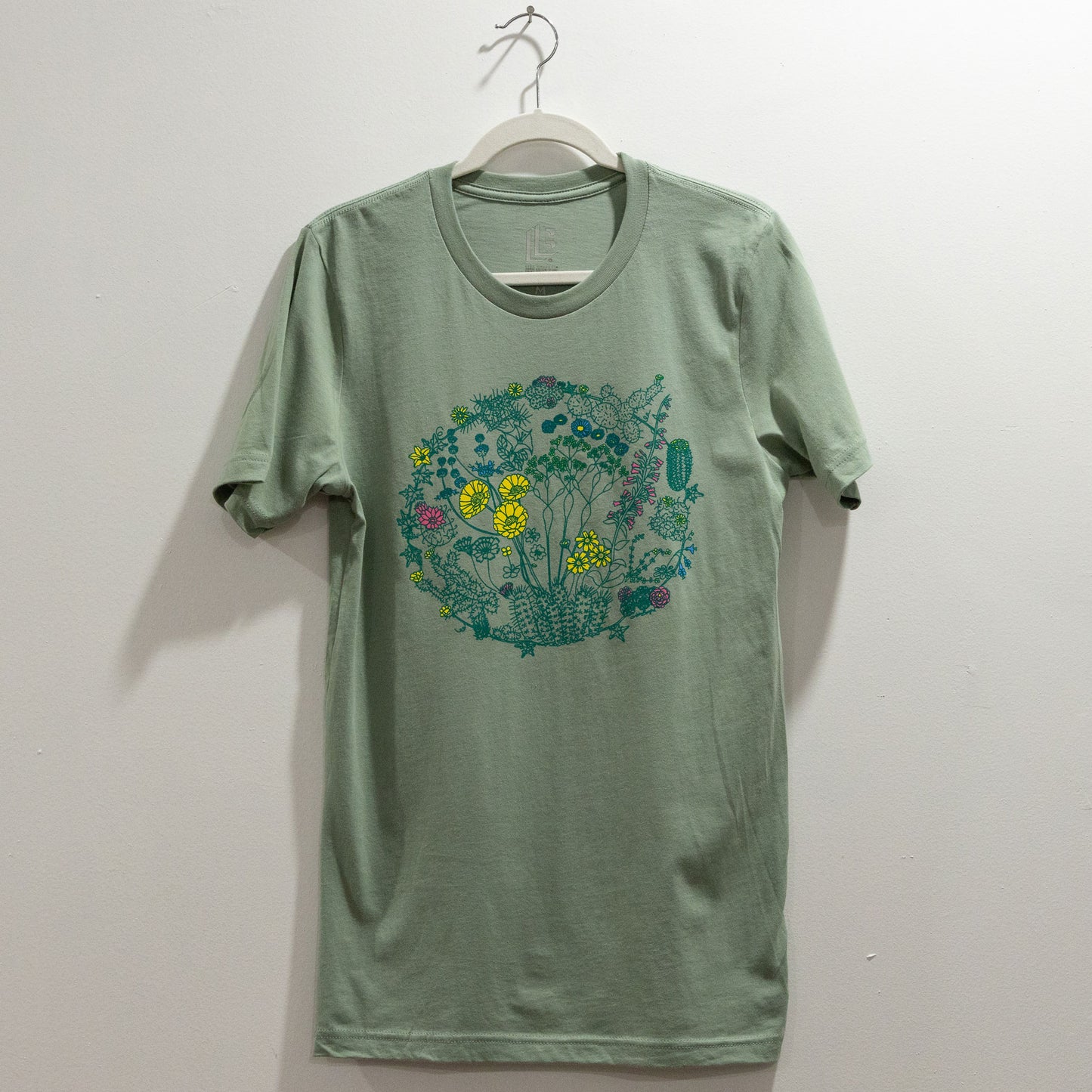 A sage color t-shirt on a hanger against a white wall. The t-shirt has a screen print of an illustration of flowers on plants that can be found in the Mojave Desert and/or Joshua Tree National Park. The outlines of the plants are a dark green color. Some of the flowers are colored a bright yellow, some a bright pink, and some a bright blue. The composition is in a circular shape.