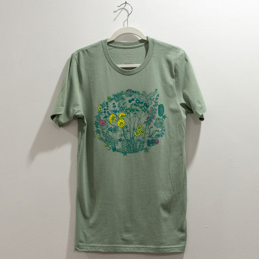 A sage color t-shirt on a hanger against a white wall. The t-shirt has a screen print of an illustration of flowers on plants that can be found in the Mojave Desert and/or Joshua Tree National Park. The outlines of the plants are a dark green color. Some of the flowers are colored a bright yellow, some a bright pink, and some a bright blue. The composition is in a circular shape.