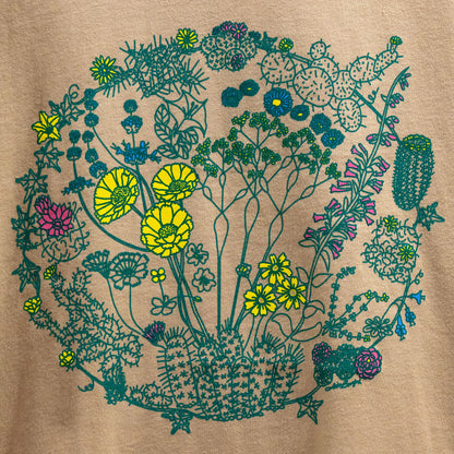 A close up of a sand color t-shirt. The t-shirt has a screen print of an illustration of flowers on plants that can be found in the Mojave Desert and/or Joshua Tree National Park. The outlines of the plants are a dark green color. Some of the flowers are colored a bright yellow, some a bright pink, and some a bright blue. The composition is in a circular shape.
