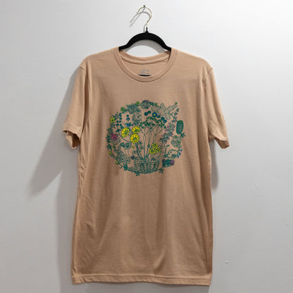 A sand color t-shirt on a hanger against a white wall. The t-shirt has a screen print of an illustration of flowers on plants that can be found in the Mojave Desert and/or Joshua Tree National Park. The outlines of the plants are a dark green color. Some of the flowers are colored a bright yellow, some a bright pink, and some a bright blue. The composition is in a circular shape.