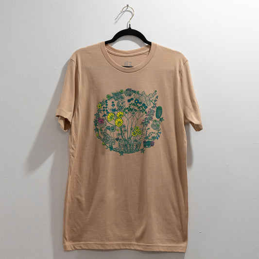 A sand color t-shirt on a hanger against a white wall. The t-shirt has a screen print of an illustration of flowers on plants that can be found in the Mojave Desert and/or Joshua Tree National Park. The outlines of the plants are a dark green color. Some of the flowers are colored a bright yellow, some a bright pink, and some a bright blue. The composition is in a circular shape.
