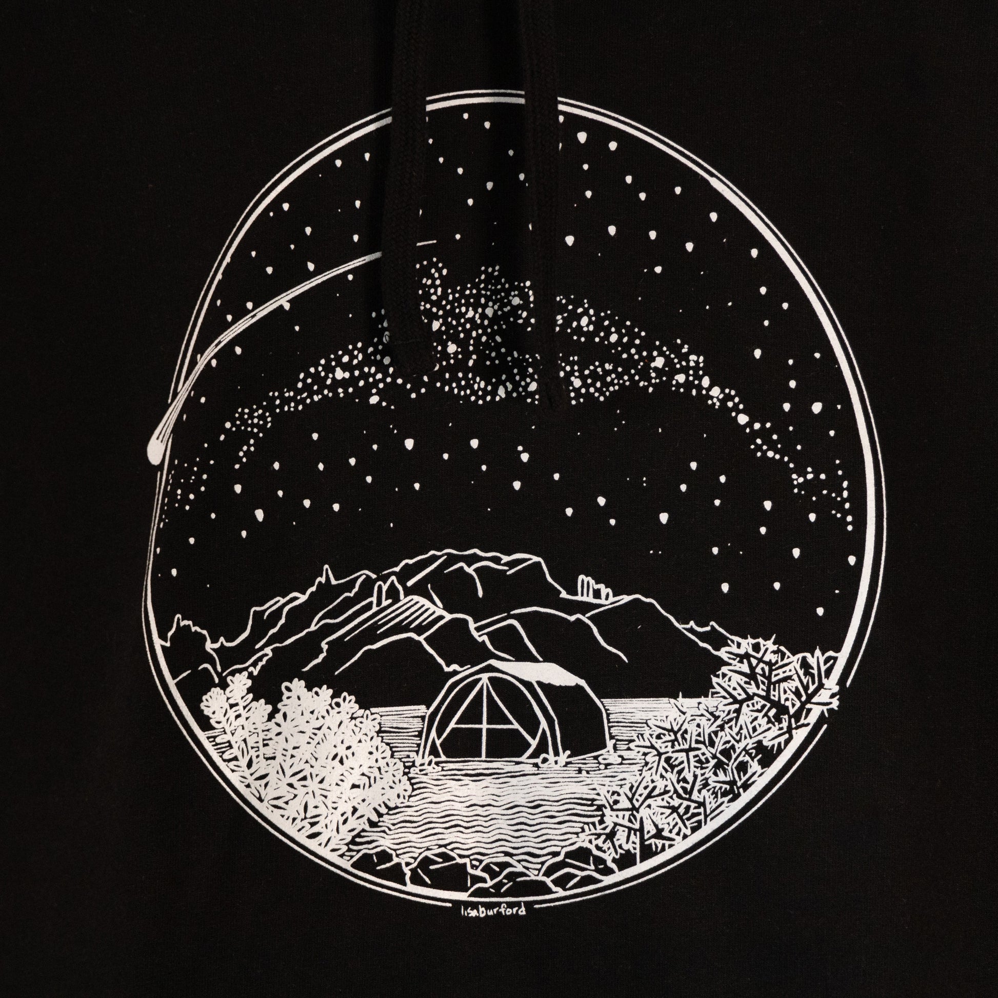 A close up of the design found on the hoodie. A white Desert Camping illustration on the front of the gender neutral black colored hooded sweatshirt. The illustration shows a tent in the foreground with mountains in the background, desert plants in the very foreground, and stars, the Milky Way gallaxy, and a shooting star in the sky above the tent. The design is encompassed in a circular shape.
