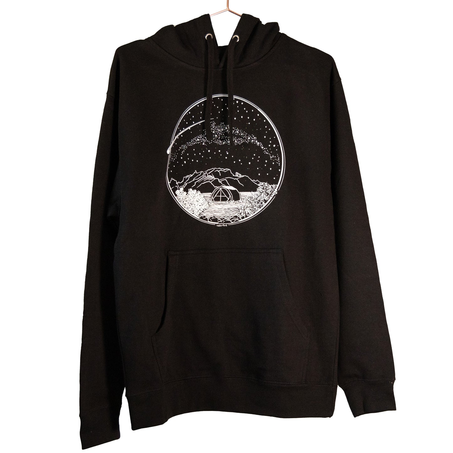 A white Desert Camping illustration on the front of the gender neutral black colored hooded sweatshirt. The illustration shows a tent in the foreground with mountains in the background, desert plants in the very foreground, and stars, the Milky Way gallaxy, and a shooting star in the sky above the tent. The design is encompassed in a circular shape.