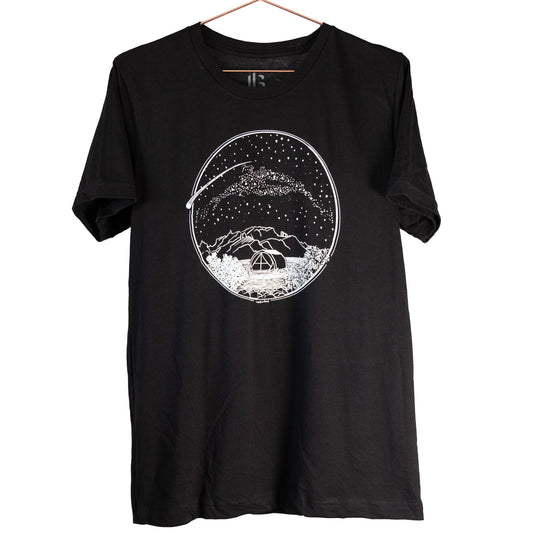 A white Desert Camping illustration on the front of the gender neutral black colored t-shirt. The illustration shows a tent in the foreground with mountains in the background, desert plants in the very foreground, and stars, the Milky Way gallaxy, and a shooting star in the sky above the tent. The design is encompassed in a circular shape.