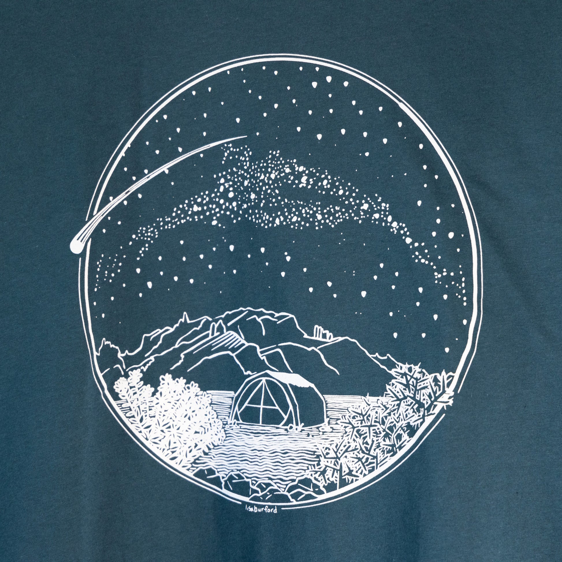 A close up of the design. A white Desert Camping illustration on the front of the gender neutral ocean blue colored t-shirt. The illustration shows a tent in the foreground with mountains in the background, desert plants in the very foreground, and stars, the Milky Way gallaxy, and a shooting star in the sky above the tent. The design is encompassed in a circular shape.