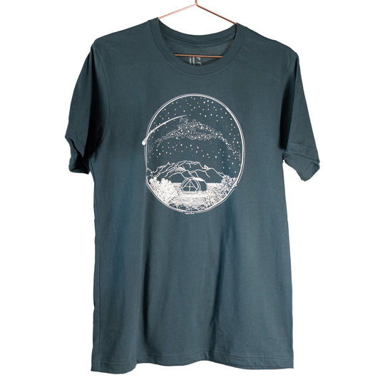 A white Desert Camping illustration on the front of the gender neutral ocean blue colored t-shirt. The illustration shows a tent in the foreground with mountains in the background, desert plants in the very foreground, and stars, the Milky Way gallaxy, and a shooting star in the sky above the tent. The design is encompassed in a circular shape.