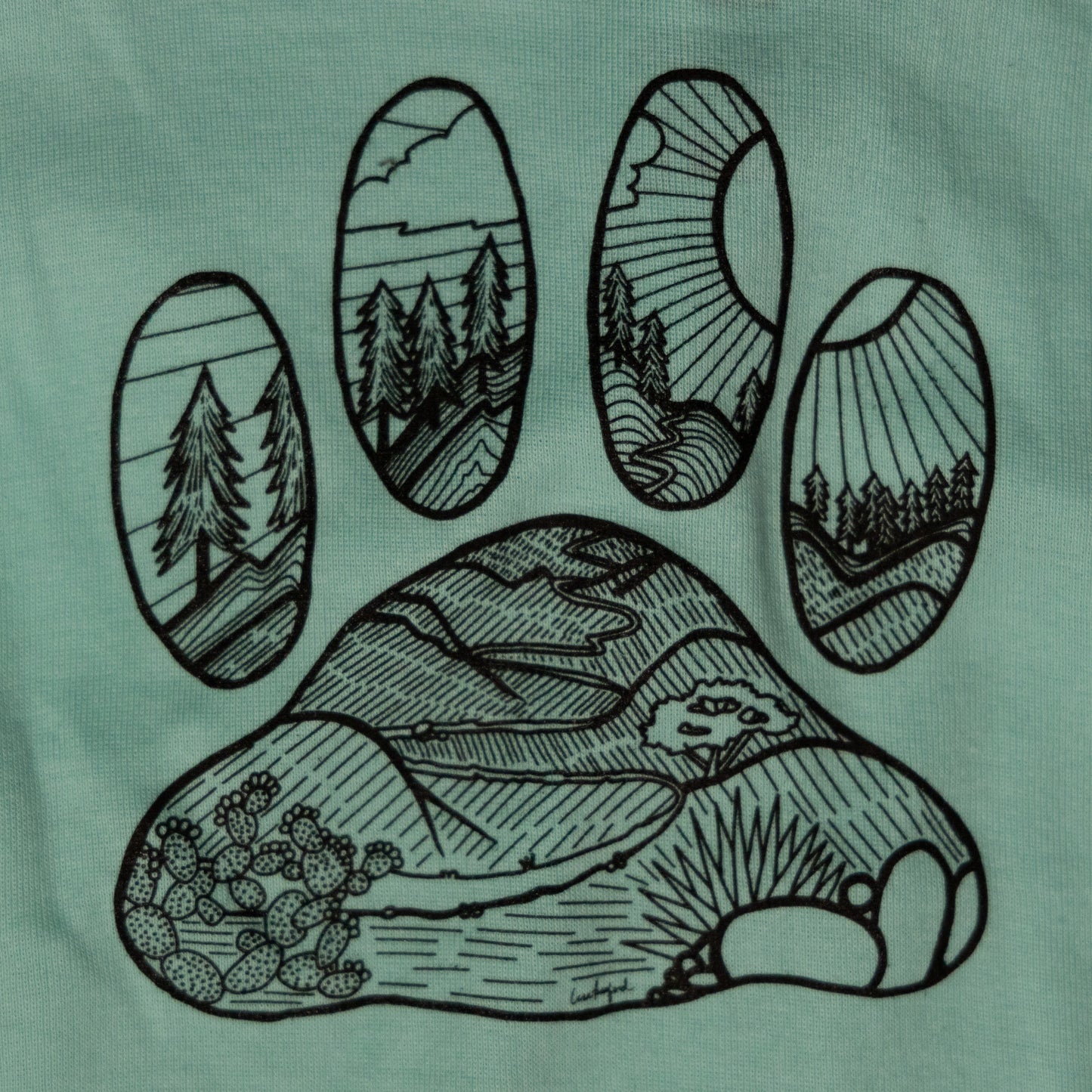 Close up of the illustrated design on the back that features a desert floor leading up to mountains with pine trees. The design is inside the shape of a dog paw print. The illustration is in black and the shirt color is light blue.