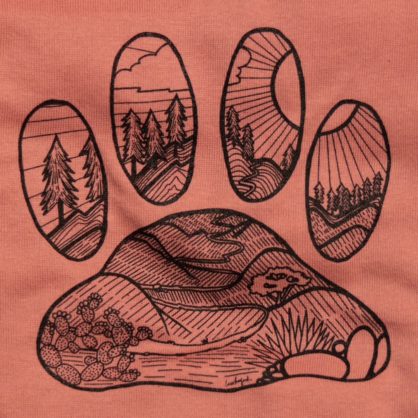 Close up of the illustrated design on the back that features a desert floor leading up to mountains with pine trees. The design is inside the shape of a dog paw print. The illustration is in black and the shirt color is mauve.