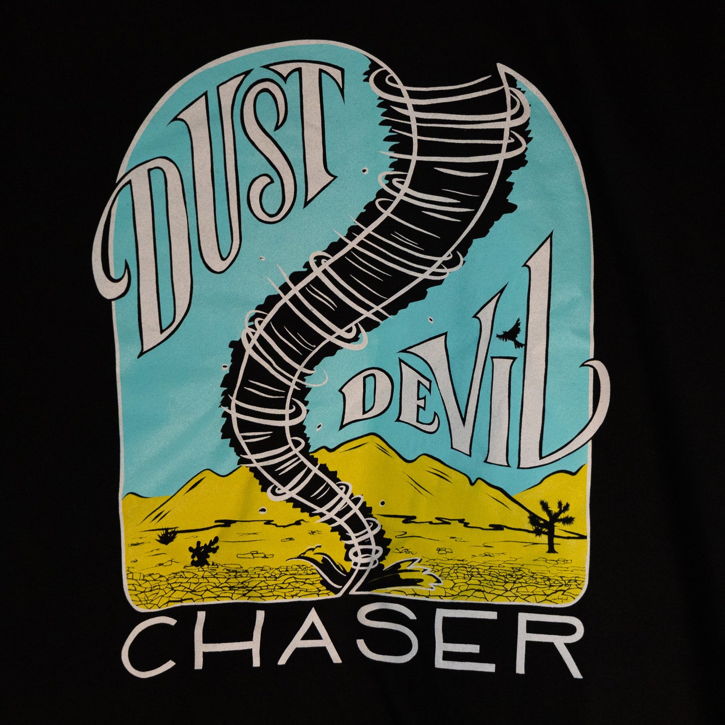 A close up of the design. The gender neutral hooded black colored t-shirt has a white, bright blue, and yellow dust devil illustration in the center. The words "Dust Devil Chaser" are written in a white handwritten font within and below the design.
