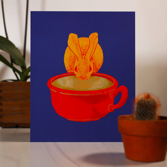 An illustrated drawing called Fika Bunny as an 8x10 inch paper print. The illustration shows a yellow furred Desert Cottontail Rabbit taking a sip out of a large mug of coffee. The coffee mug is red in color and has a handle on the right side. The background of the illustration is a bright blue.