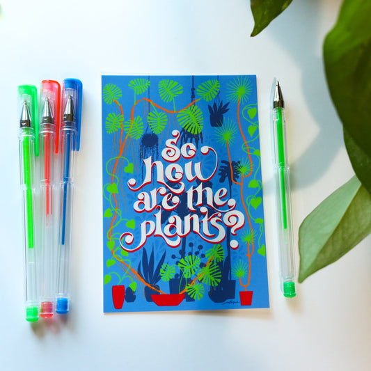 An illustrated print with the hand lettered question "So how are the plants?" and various plants surrounding the letters from the ground up or hanging overhead. Next to the print are 4 gel pens and pothos leaves on the right side.
